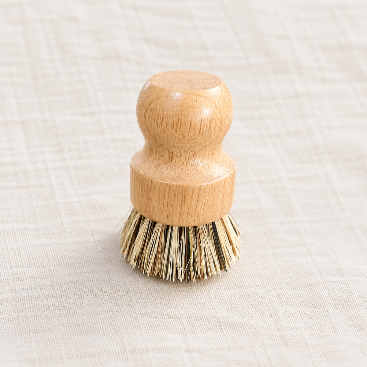 A small wooden-handled cleaning brush with natural bristles, photographed on a light beige textured fabric background.