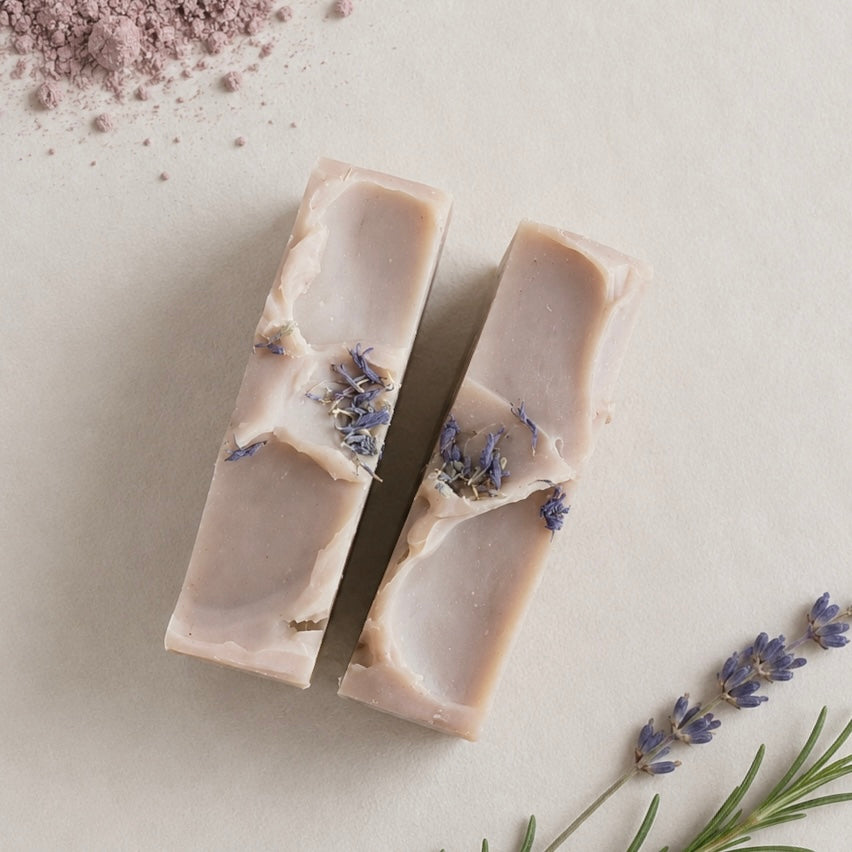 Two rectangular bars of pale lavender soap with dried lavender buds embedded inside, arranged on a light beige surface with scattered lavender sprigs and powder.