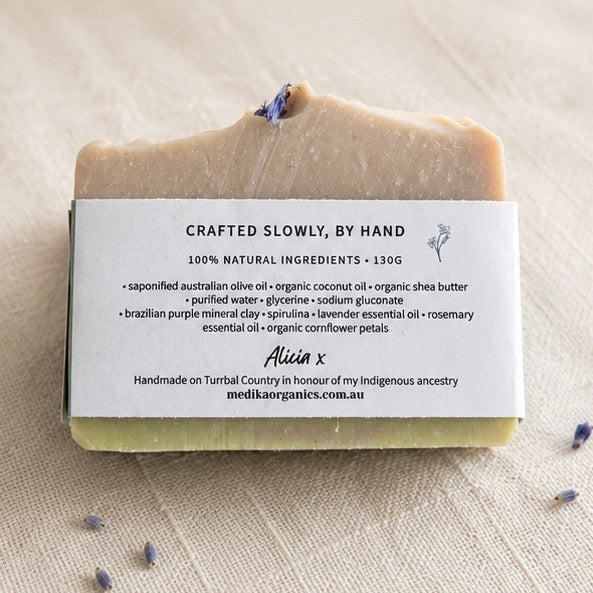 A handmade bar of soap with a rustic, uneven top, featuring lavender buds on top. The soap has a beige top layer and a pale green bottom layer, wrapped in a white paper label that reads 'CRAFTED SLOWLY, BY HAND' and lists ingredients.