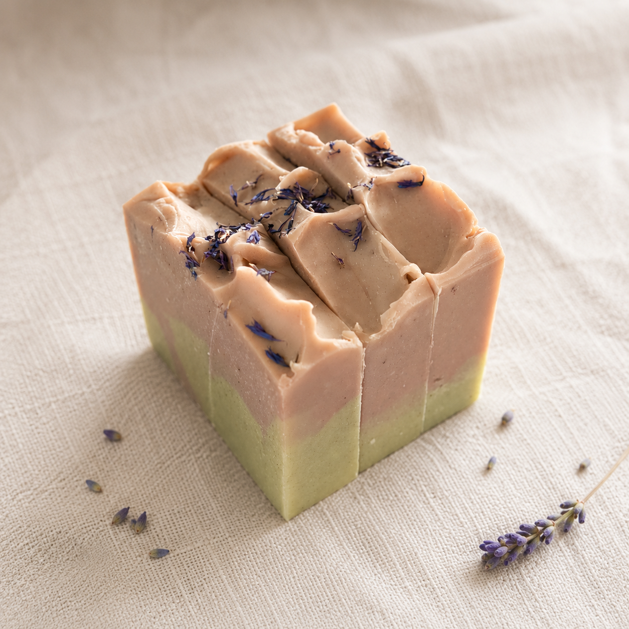 Three layered handmade soap bars with a light pink top layer and a pale green bottom layer, decorated with dried lavender buds, set on a textured beige linen fabric background.