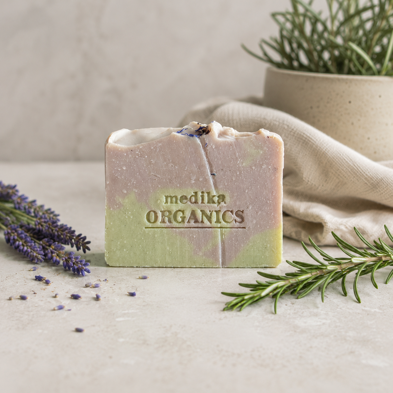 A rectangular handmade soap bar with a marbled lavender and light green color, embossed with the brand name 'medika ORGANICS'. The soap is styled on a light stone surface with sprigs of lavender and rosemary, a beige linen cloth, and a potted plant in the background.