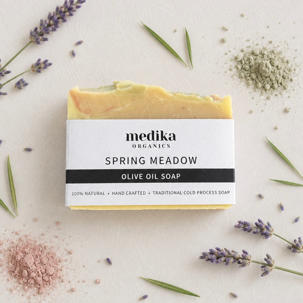 A rectangular bar of Medika Organics Spring Meadow Olive Oil Soap is centered on a light textured background, surrounded by lavender sprigs, green leaves, and small piles of dried botanical powders. The soap has a white paper label with black text and shows a pale yellow-green, peach, and cream marbled soap bar through the top.