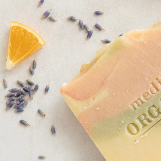 A close-up of a handmade soap bar with peach, yellow, and pale green marbled layers, embossed with the text 'Medika Organics'. A slice of orange and scattered lavender buds are styled on a light neutral background.