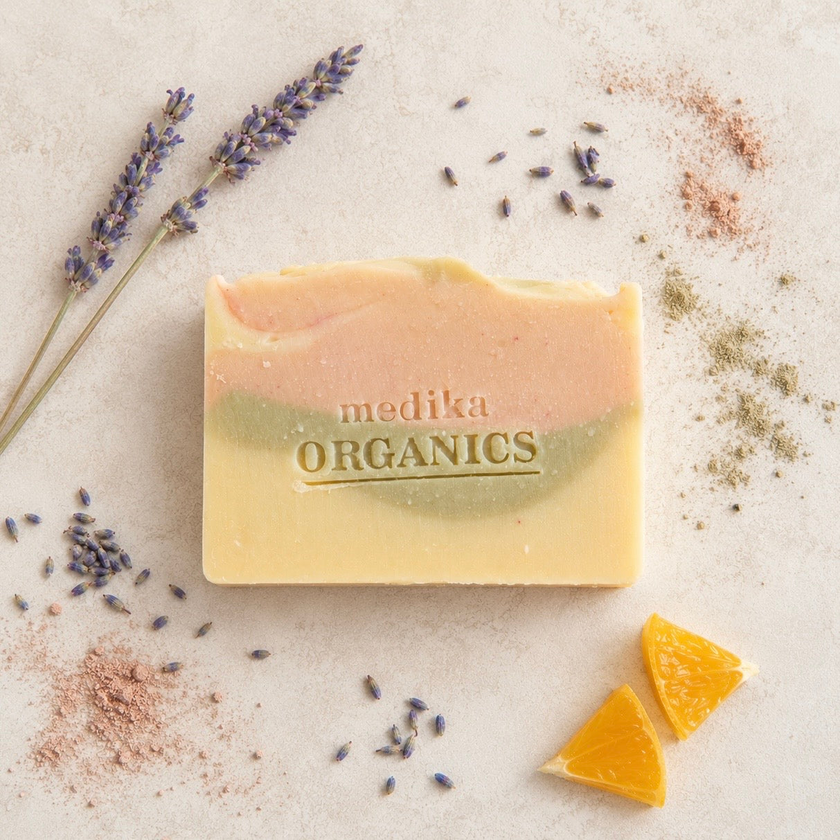 A rectangular bar of Medika Organics soap with a three-color swirled design of pale yellow, peach, and sage green, styled on a textured light beige surface with lavender sprigs, orange slices, and botanical powders.