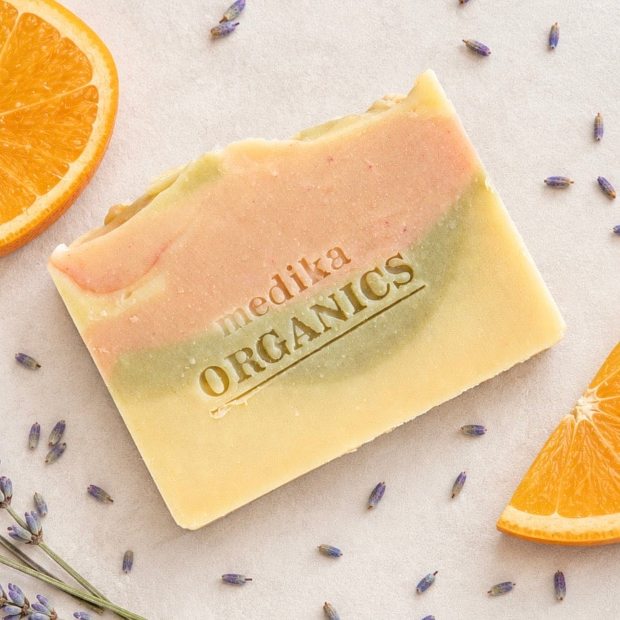 A rectangular bar of handmade soap with a swirled pastel design in pink, green, and pale yellow, embossed with the brand name 'Medika Organics'. The soap is styled on a light beige surface with scattered lavender buds, orange slices, and lavender sprigs.