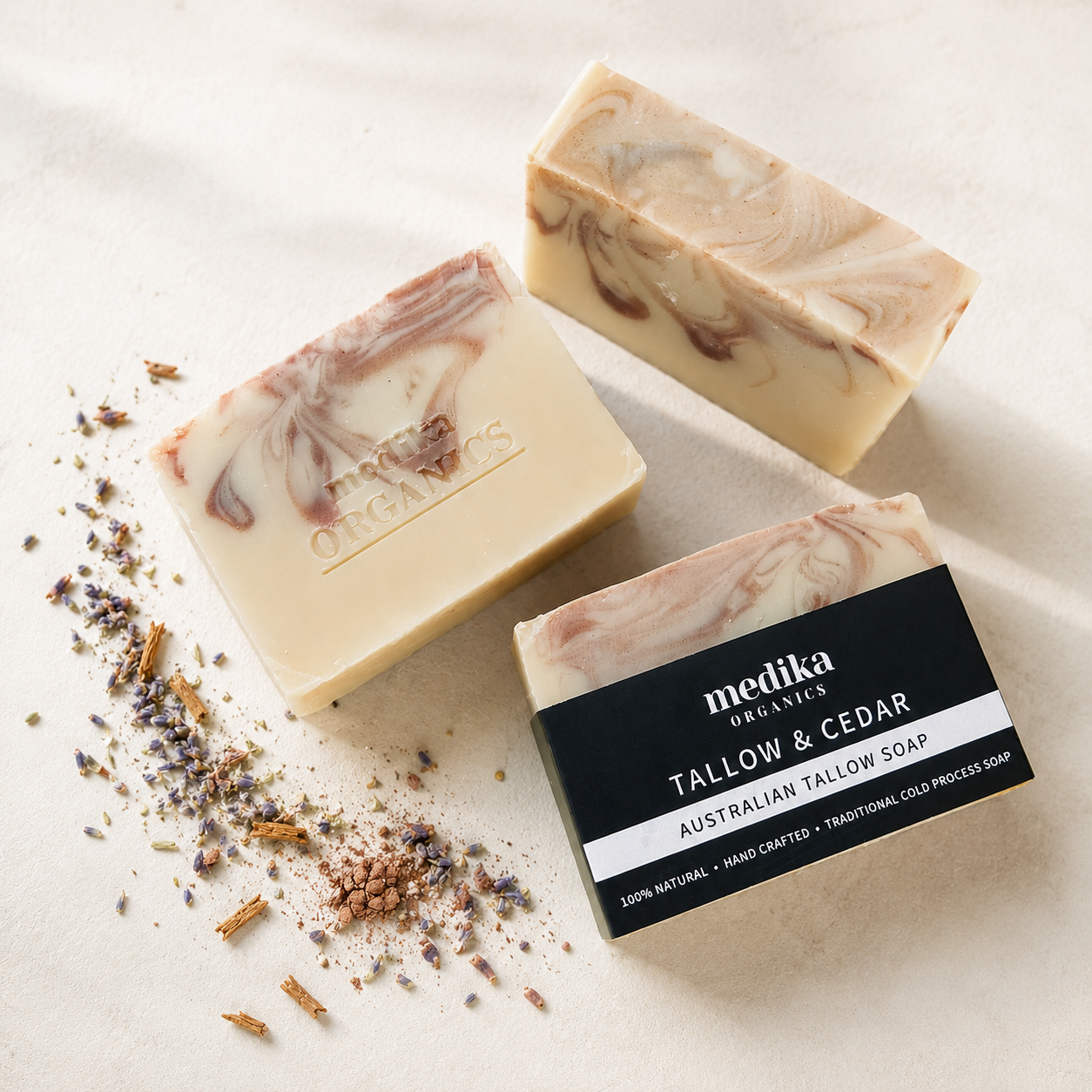 Three bars of Metika Organics Australian Tallow & Cedar soap are arranged on a light beige textured surface with scattered dried herbs. One bar is shown with its black and white retail label, while the other two are bare.