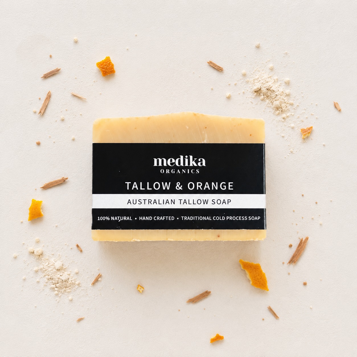 A rectangular bar of Medika Organics Tallow & Orange Australian Tallow Soap, featuring a black label with white text, styled on a light beige background with scattered orange peel and wax shavings.