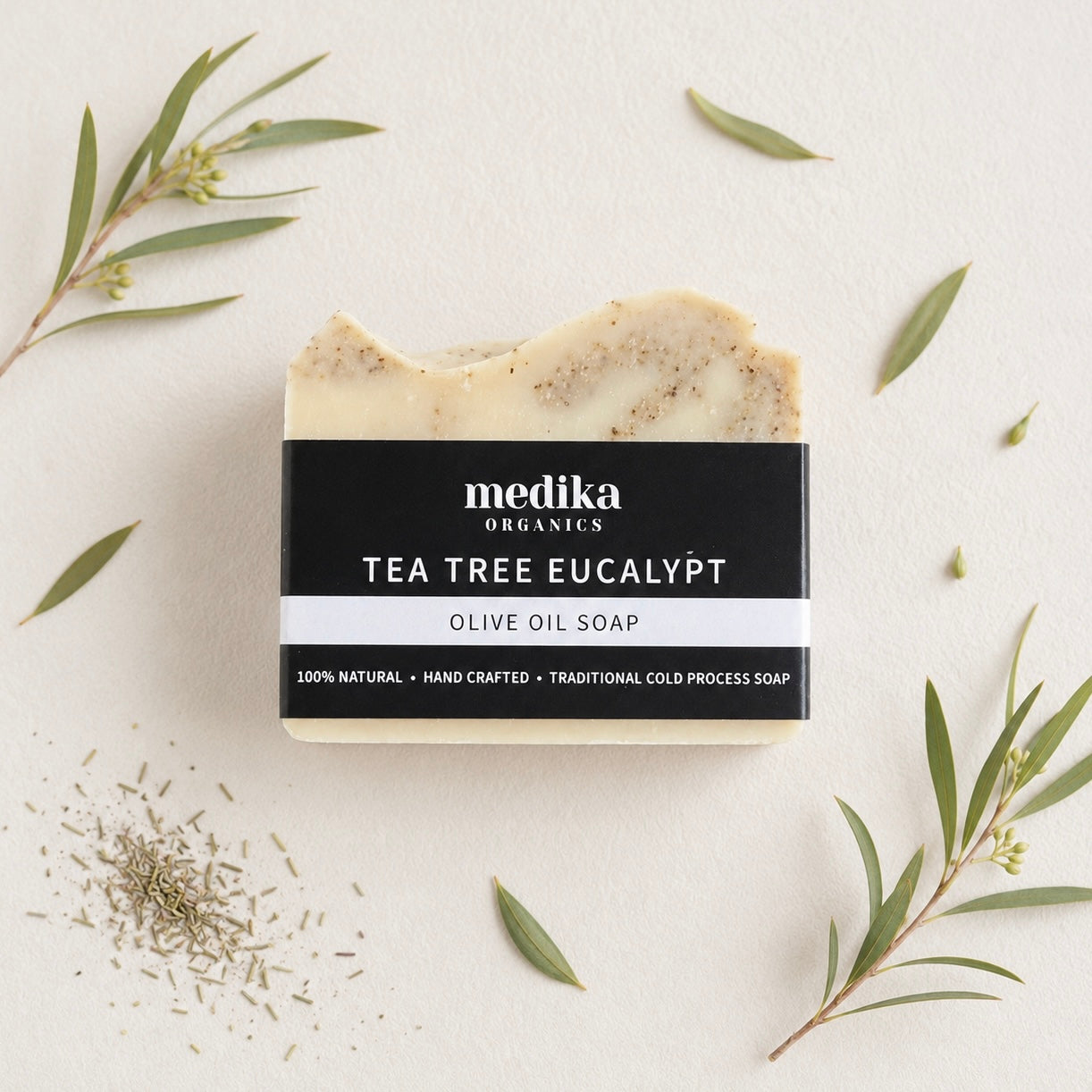 A bar of Medika Organics Tea Tree Eucalyptus Olive Oil Soap is shown in a styled flat-lay arrangement with olive branches and loose tea tree leaves on a textured off-white background.