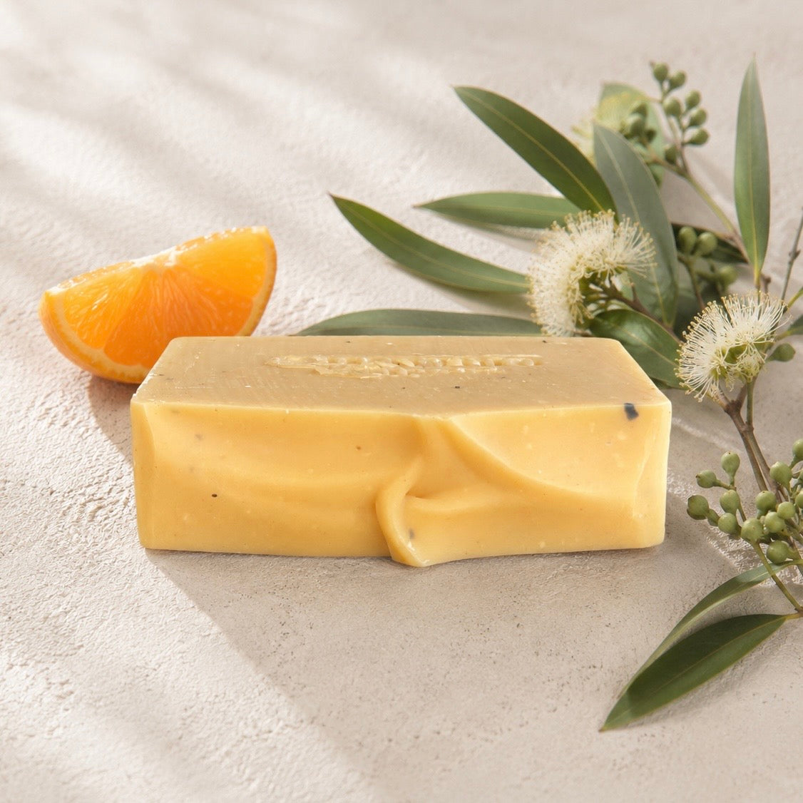 A rectangular bar of yellow soap with a textured surface and embossed with Medika Organics logo, styled on a light beige textured surface with an orange slice, green leaves, and white flowers.