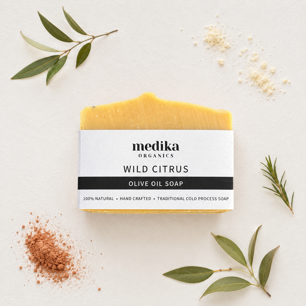 A bar of Medika Organics Wild Citrus Olive Oil Soap is displayed on a textured off-white background, styled with olive leaves, rosemary sprigs, and spices. 