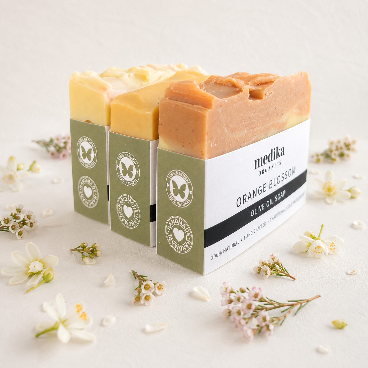 Three bars of Medika Organics Olive Oil Soap in Orange Blossom scent, displayed in their green and white packaging with small white flowers scattered around on a light beige background.
